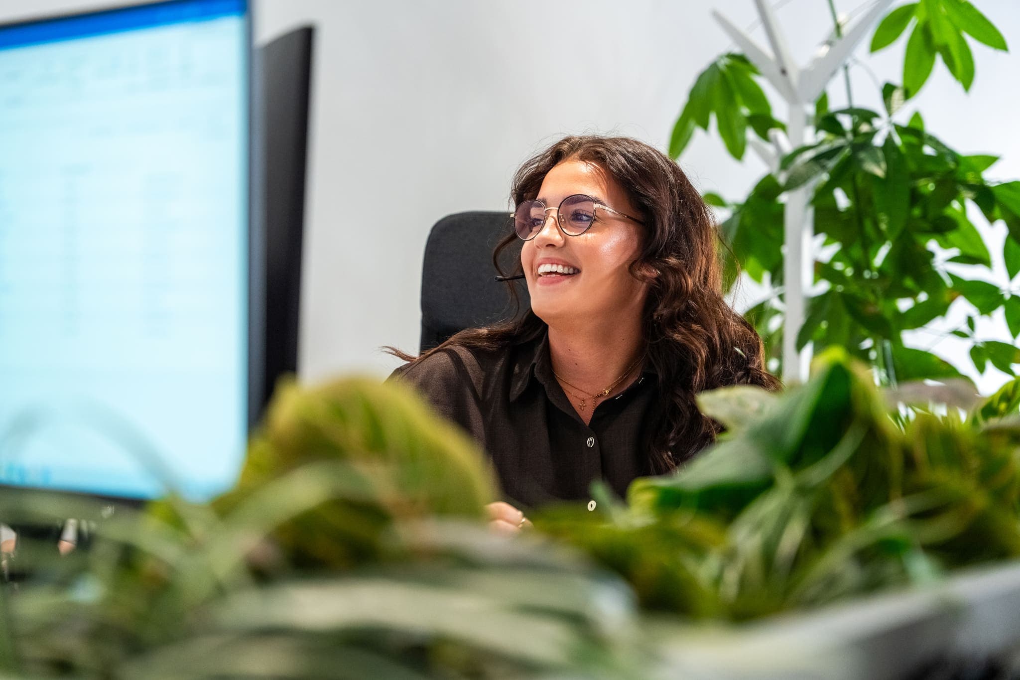 Vrouw met bril lacht achter een bureau tussen planten, ConnectYou