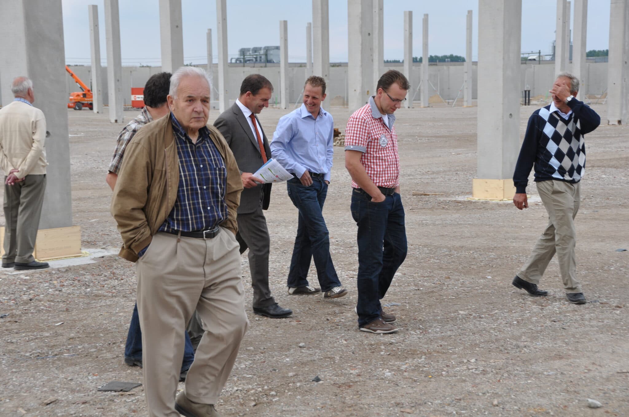 Groep mannen loopt over een bouwterrein met betonnen kolommen, voor Plantion