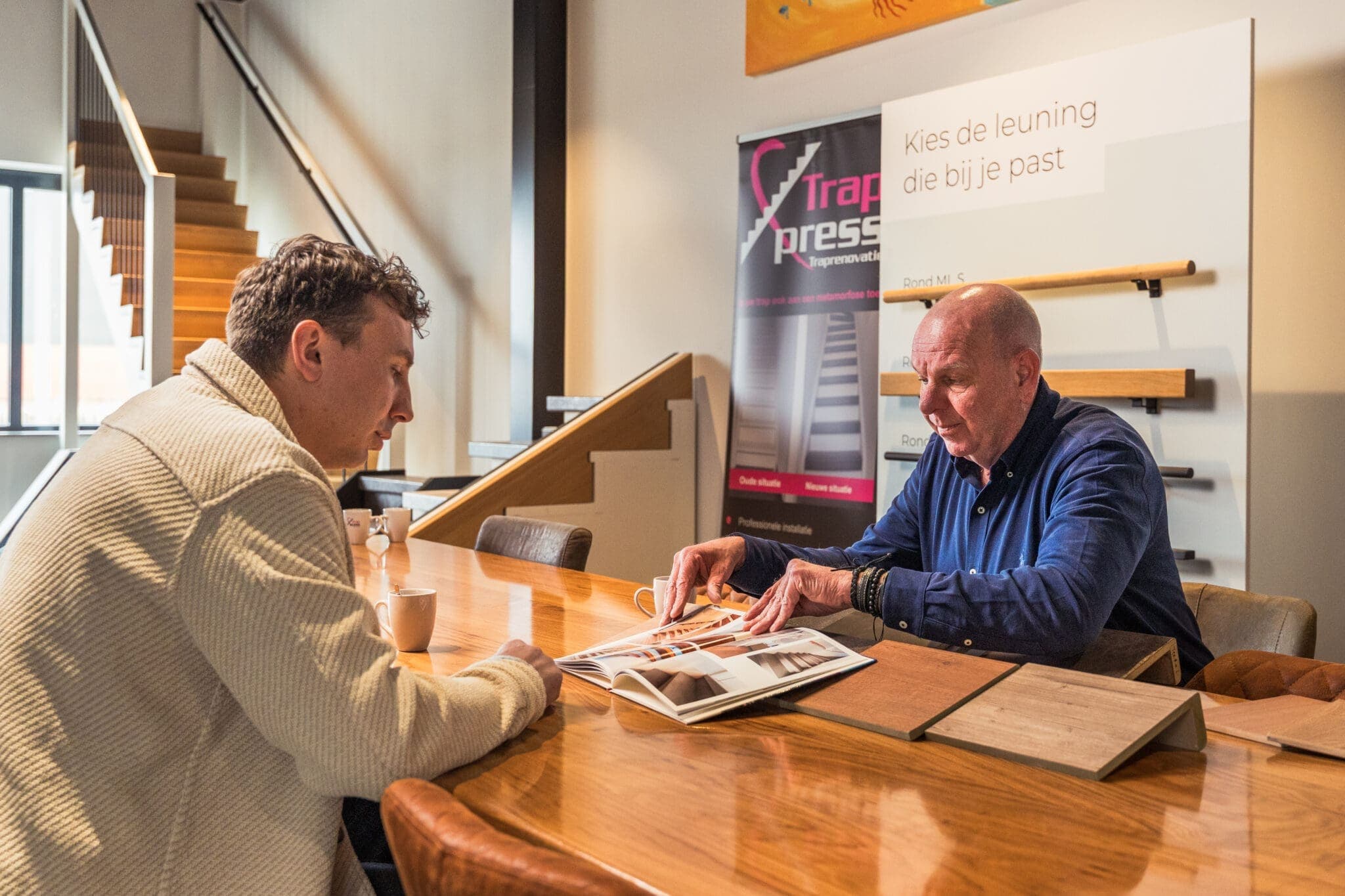 Twee mannen bekijken een brochure met houtstalen aan tafel in de showroom van TrapXpress