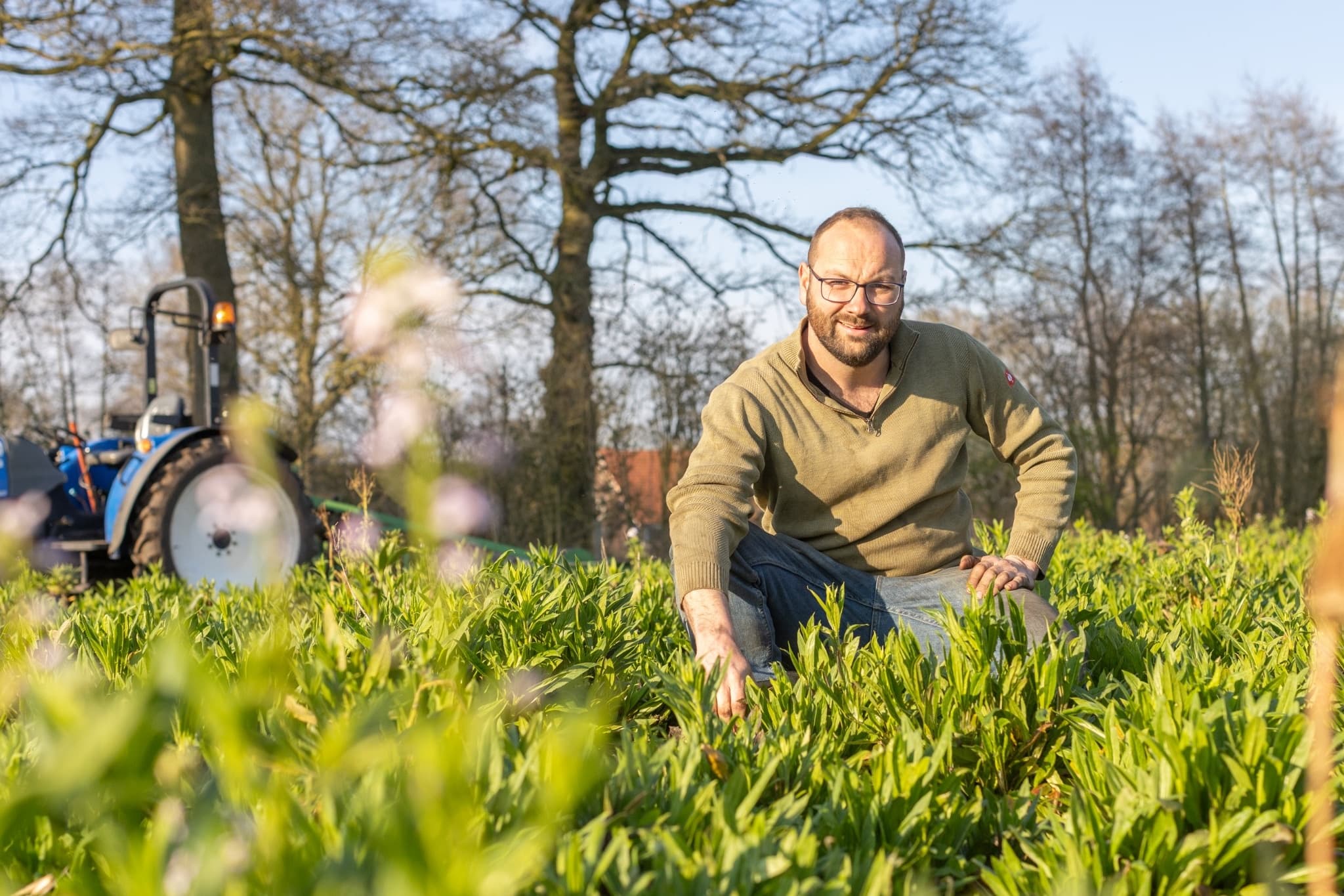Man met bril hurkt tussen jonge planten op een veld met een tractor op de achtergrond, voor Plantion